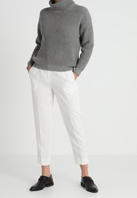 Grauer gestrickter Rollkragenpullover mit strukturiertem Muster, kombiniert mit weißen maßgeschneiderten Hosen und schwarzen Slippern.