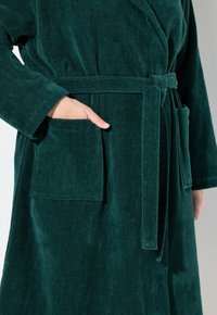 Accappatoio in corduroy verde con cintura, tasche laterali e una texture morbida. Il design presenta maniche lunghe e una linea dritta.