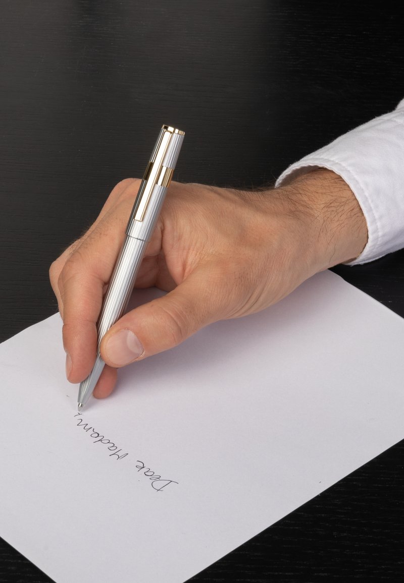 Main droite tenant un stylo argenté avec des accents dorés écrivant "Chère Madame" sur du papier blanc posé sur une surface noire.