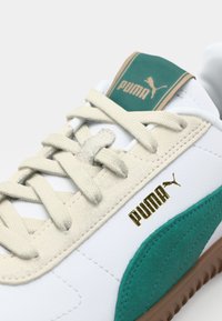Valged tennisjalanikud rohelise siidiga detailidega, beežide paeltega ja kuldse "PUMA" logoga küljel. Keskendub mugavusele ja juhuslikule stiilile.