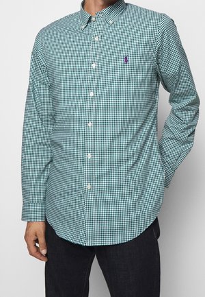 Homme portant une chemise à manches longues à carreaux verts et blancs avec un petit logo brodé violet sur la poitrine.
