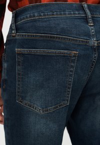 Gros plan sur la poche arrière d'un jean en denim bleu avec des coutures orange-brun, porté avec une chemise à carreaux rouge.