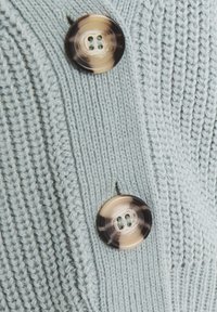 Ljusblått stickat cardigan med framträdande ribbningar och två stora, cirkulära knappar med ett virvat mönster i brunt och krämvitt.
