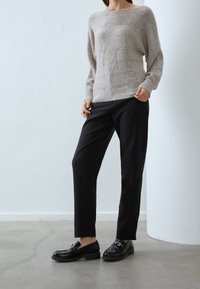 Femme portant un pull beige texturé, un pantalon noir cintré et des mocassins noirs, debout sur un sol gris contre un mur blanc.