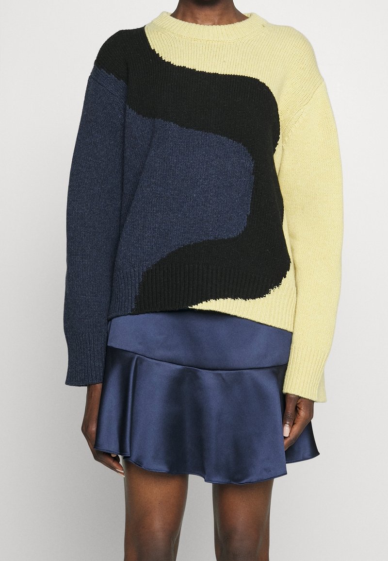 Strickpullover in Gelb, Marineblau und Schwarz mit abstraktem Muster; lockere Passform, gerippter Halsausschnitt und Ärmelbündchen; darüber getragen über einem blauen Satinskirt.