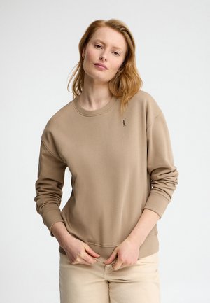 Jeune femme aux cheveux roux clairs portant un sweat-shirt beige et un pantalon crème, debout devant un fond clair uni.