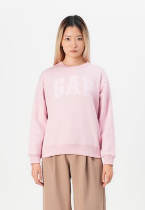 Jeune femme aux longs cheveux portant un sweat-shirt rose GAP et un pantalon beige, se tenant devant un fond blanc uni.
