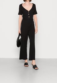 Czarny prążkowany kombinezon z krótkimi rękawami, dekoltem w kształcie V i trzema guzikami. Posiada przewiązany w talii krój i szerokie nogawki. Połączony z plecionymi czarnymi klapkami i torebką.