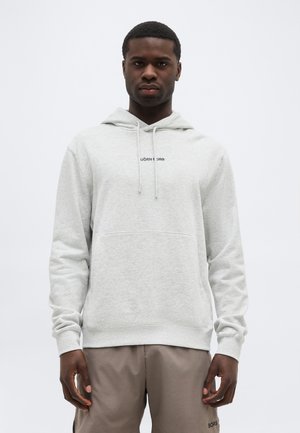 Mand iført en lysegrå hættetrøje med frontlomme og beige bukser, stående foran en ensfarvet hvid baggrund.