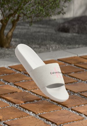 White Calvin Klein slide sandal with pink and black logo on textured outdoor wooden tile flooring near a small tree.