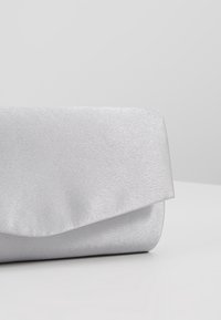 Pochette rectangulaire argentée à finition satinée, avec une fermeture à rabat plié, posée sur une surface gris clair.