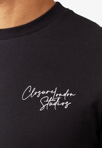 Közeli kép fekete, kerek nyakú pólóról, melyen a mellrészén fehér felirat: "Closure London Studios" szerepel.