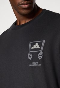 Schwarzes Baumwoll-T-Shirt mit einem grauen Druckdesign von Händen, die eine Fahne mit drei Streifen halten, und dem Text "ADIDAS SPORTSWEAR" darunter.