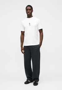 T-shirt en coton blanc avec un motif graphique noir central. Associé à un pantalon ample foncé et des chaussures de sport noires avec des accents blancs.