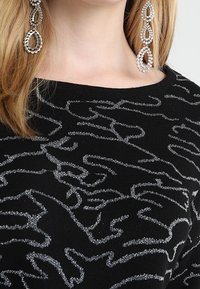 Schwarzes Strickoberteil mit einem silbernen abstrakten Muster. Der Ausschnitt ist weit und der Stoff scheint eine strukturierte Textur zu haben. Lange, verzierte Ohrringe ergänzen den Look.