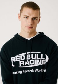 Svart hoodie med "RED BULL RACING" i fet vit text. Justerbar dragsko och mjuk textur. Klassisk passform som passar för avslappnad användning.