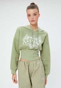 Sudadera verde recortada con capucha y cordón, texto gráfico blanco y cinta ribeteada en la cintura. Mangas de corte holgado y diseño casual. Combinada con pantalones beiges.