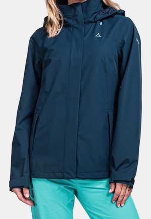 Femme portant une veste imperméable à capuche bleu marine avec fermeture éclair et un pantalon turquoise, montrée du cou aux hanches.