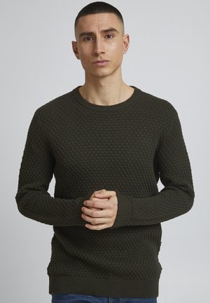 Suéter de punto verde oscuro con un patrón texturizado de panal, cuello redondo y puños y dobladillo acanalados. Modelo de pie con las manos entrelazadas.