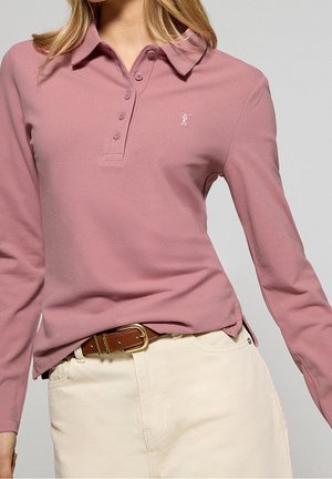 Kobieta w różowym, długim rękawem polo, włożonym do kremowych spodni z brązowym skórzanym paskiem.