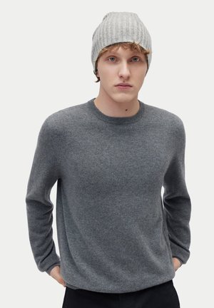 Junger Mann mit heller Haut, der eine graue Strickmütze und einen grauen Pullover mit langen Ärmeln trägt, steht mit den Händen in den Taschen vor weißem Hintergrund.