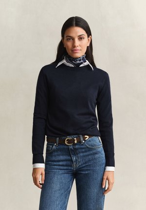 Vrouw met lang donker haar die een marineblauwe trui draagt over een wit overhemd met kraag, een patroonachtige nek sjaal, blauwe jeans en een bruine riem.