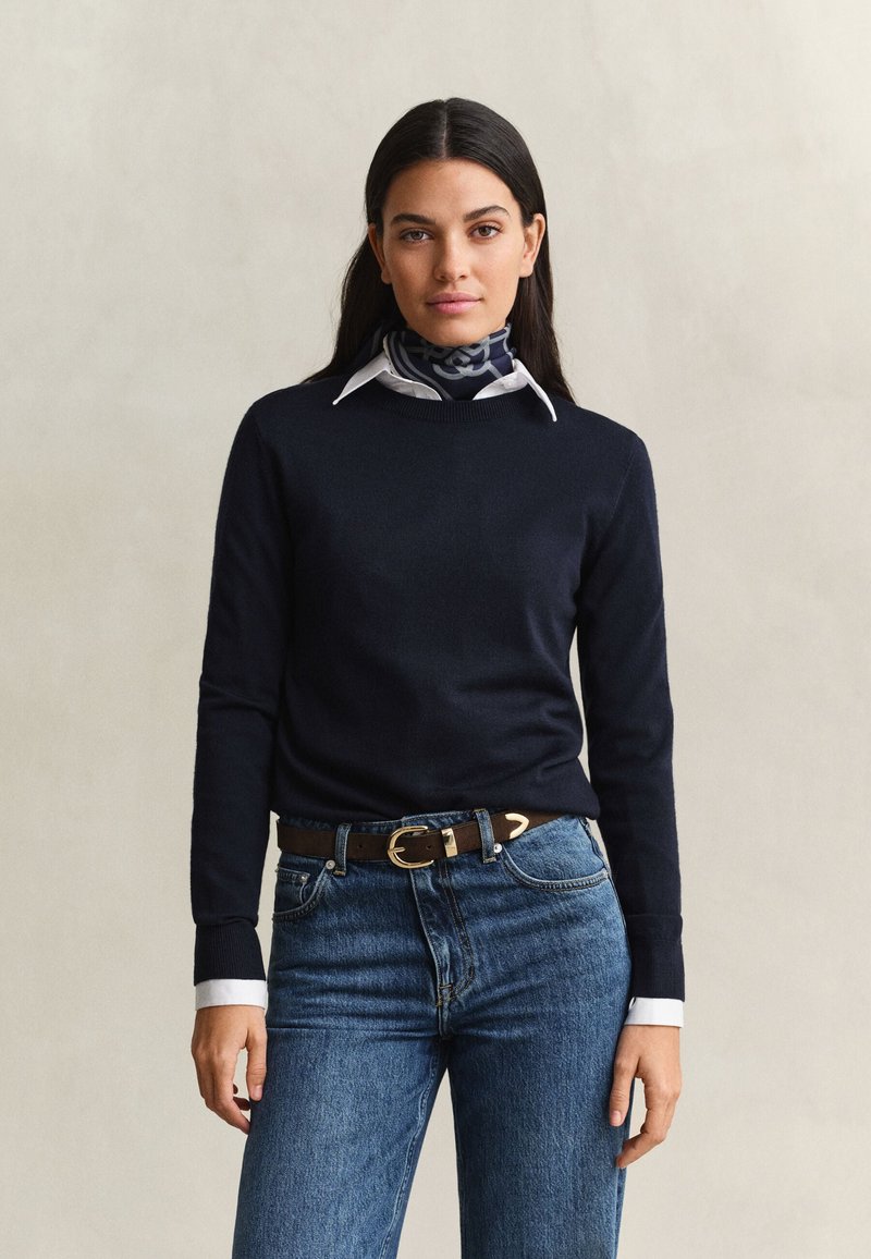 Donna con lunghi capelli scuri che indossa un maglione blu navy sopra una camicia bianca con colletto, una sciarpa fantasia al collo, jeans blu e una cintura marrone.