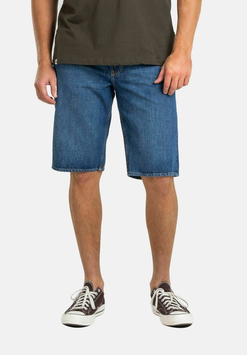 Shorts de denim en azul medio, a la altura de la rodilla, con un corte recto y dobladillo cosido. Combinados con zapatillas oscuras y una camiseta de manga corta en color oliva.