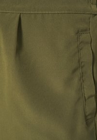 Gros plan sur un tissu vert olive montrant un pli avant et une poche latérale insérée avec des coutures visibles.