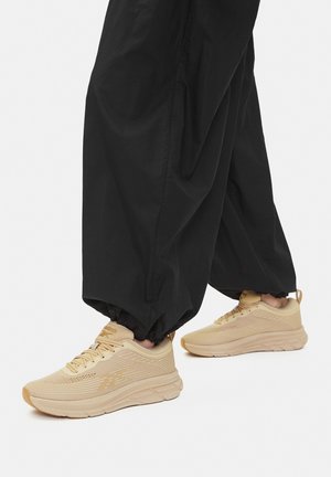 Beżowe buty sportowe z teksturowaną siateczkową cholewką i systemem sznurowania, połączone z luźnymi czarnymi spodniami, ukazujące kontrast kolorów i stylów.