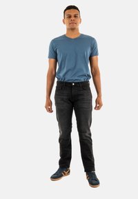 Camiseta azul de manga corta con escote en V, jeans oscuros y zapatillas azules con acentos naranjas, de pie sobre un fondo neutro.