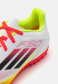Botas de fútbol blancas con una superficie texturizada, que presentan acentos de un amarillo brillante, cordones rojos y un diseño a rayas negro y rosa.