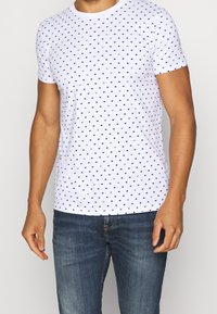 Hombre vistiendo una camiseta blanca ajustada con un pequeño patrón de banderas azules y jeans oscuros, de pie con los brazos relajados a los lados.