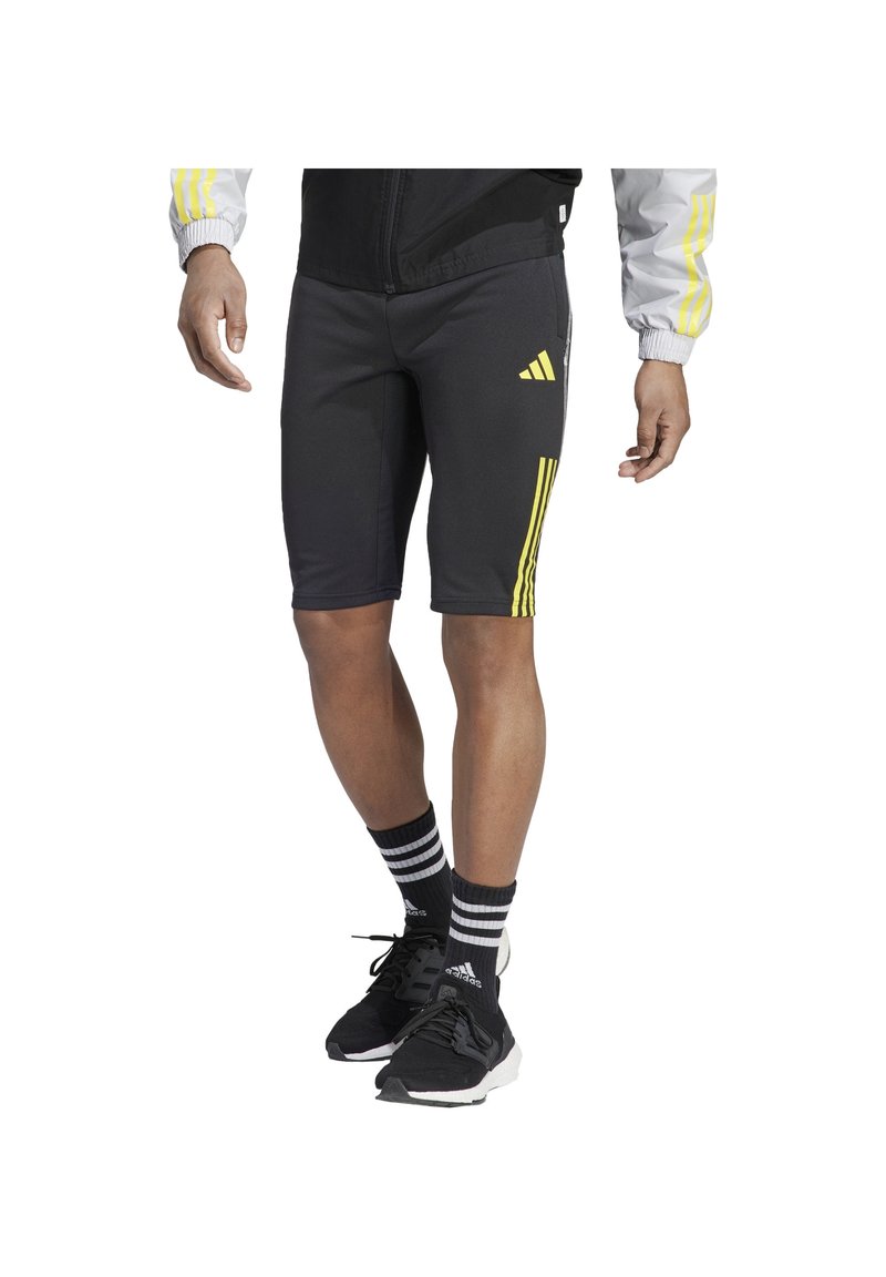 Homme portant un short de sport noir avec le logo et les bandes jaunes Adidas, des chaussettes noires Adidas, des baskets noires et une veste grise avec des bandes jaunes.