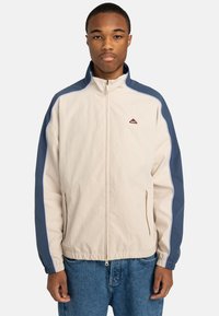 Light beige zip-up jacket with blue side panels, elastic cuffs, two zippered pockets, and a small logo on the chest.