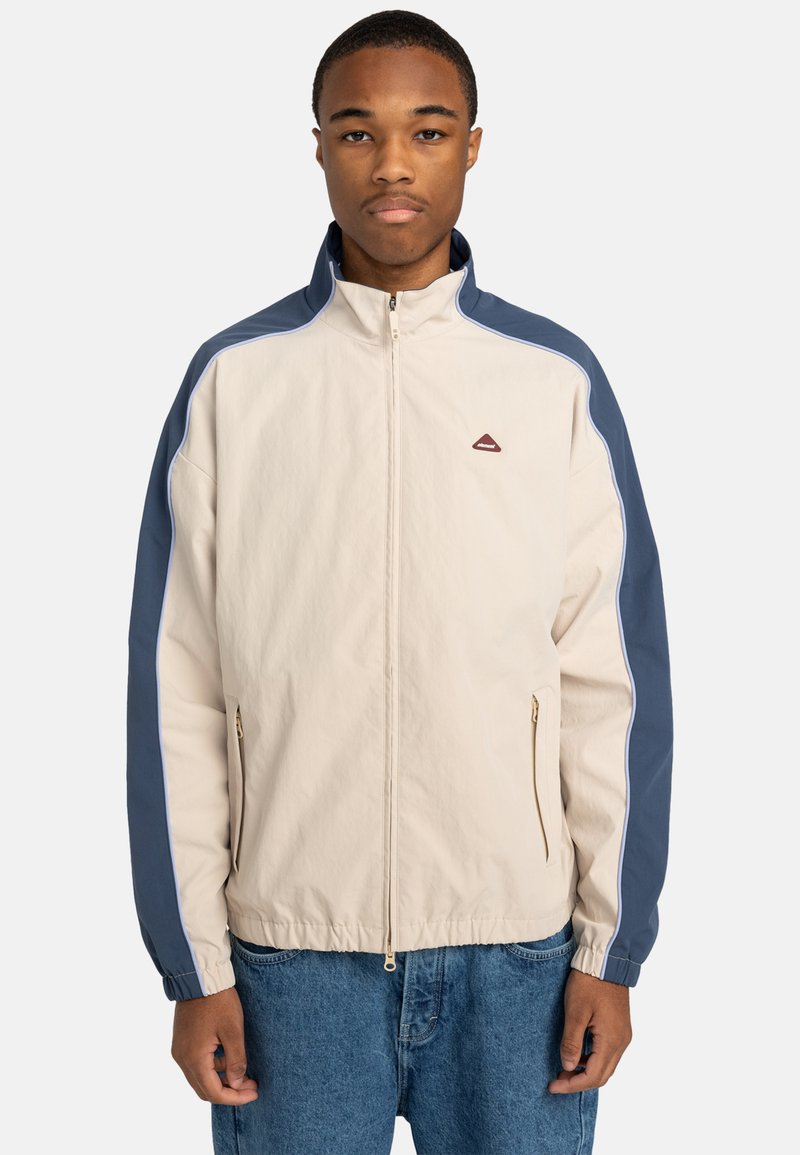 Light beige zip-up jacket with blue side panels, elastic cuffs, two zippered pockets, and a small logo on the chest.