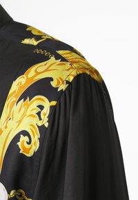 Zwarte shirt met gele sierlijke patronen op de schouder. De stof lijkt glad met een lichte glans, en de details rondom de naad zijn zichtbaar.