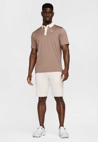 Brūns polo krekls ar bēša krāsas apkakli, ar attēlu uz krūtīm. Kombinēts ar baltām īsām biksēm un baltām sporta kurpēm. Modelis stāv pret vienkāršu fonu.