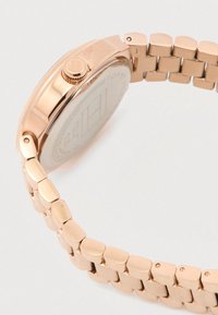 Rozā zelta metāla pulkstenis ar saistītu aproci un regulēšanas kroni, attēlots no sāna, koncentrējoties uz aizmugures un bandas detaļām.