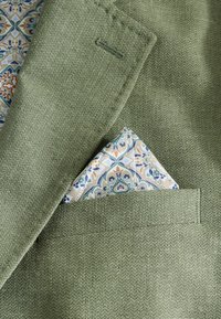 Grüner strukturierter Stoff mit einem Taschentuch, das ein florales Muster in Blau- und Orangetönen aufweist und in eine Brusttasche gefaltet ist.