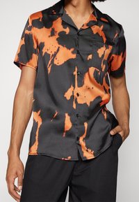 Kurzarmhemd in einem schwarz-orangenen Batikmuster, aus glattem, glänzendem Stoff; mit Knopfleiste und Brusttasche.