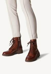 Bottes en cuir marron à lacets avec une fermeture éclair sur le côté, à bout rond et semelle en caoutchouc texturé, associées à un pantalon slim clair.