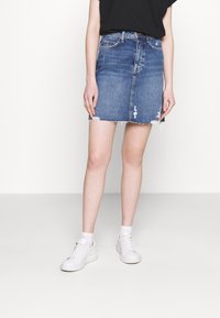 Jeansrok met een rafelige zoom, lichtblauwe was en versleten plekken. Draag het met een zwart topje en witte sneakers, staande voor een effen achtergrond.