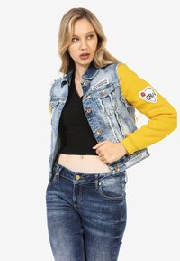 Veste en denim avec des manches jaunes, comportant un effet vieilli, des boutons, des patchs et une coupe courte, assortie à un jean en denim bleu foncé.