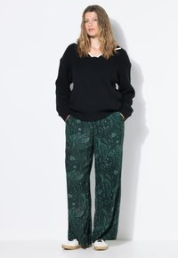 Femme aux cheveux ondulés portant un pull en tricot noir, un pantalon large à motifs verts et des baskets blanches, se tenant avec les mains dans les poches.