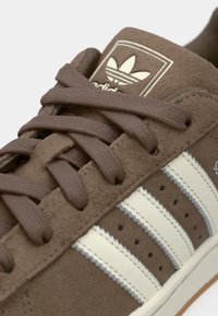 Sneaker in suede marrone con tre strisce bianche, lacci marroni e punta arrotondata. Include il logo Adidas su una linguetta imbottita.