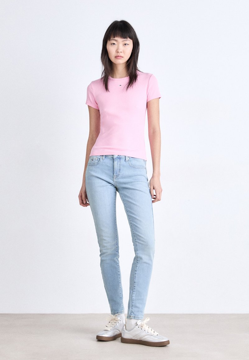 Rosa Baumwoll-T-Shirt mit kurzen Ärmeln, kombiniert mit hellblauen Skinny-Jeans und silbernen Turnschuhen mit weißen Sohlen vor einem neutralen Hintergrund.