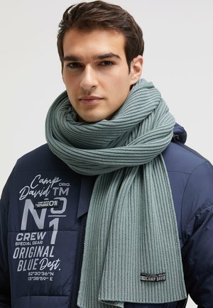 Hellblau-grün gestreifter Strickschal, der um den Hals drapiert ist, kombiniert mit einer blauen Pufferjacke, die bedruckte Marken- und Aufnähpatches aufweist.