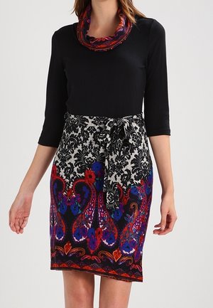 Robe noire à manches longues avec une jupe à motif floral dans les tons rouge, bleu et violet, ceinturée à la taille avec une écharpe assortie.