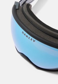Ochelari de schi Oakley cu tentă albastră, umplutură din spumă și ramă neagră, în fața unui fundal alb simplu.